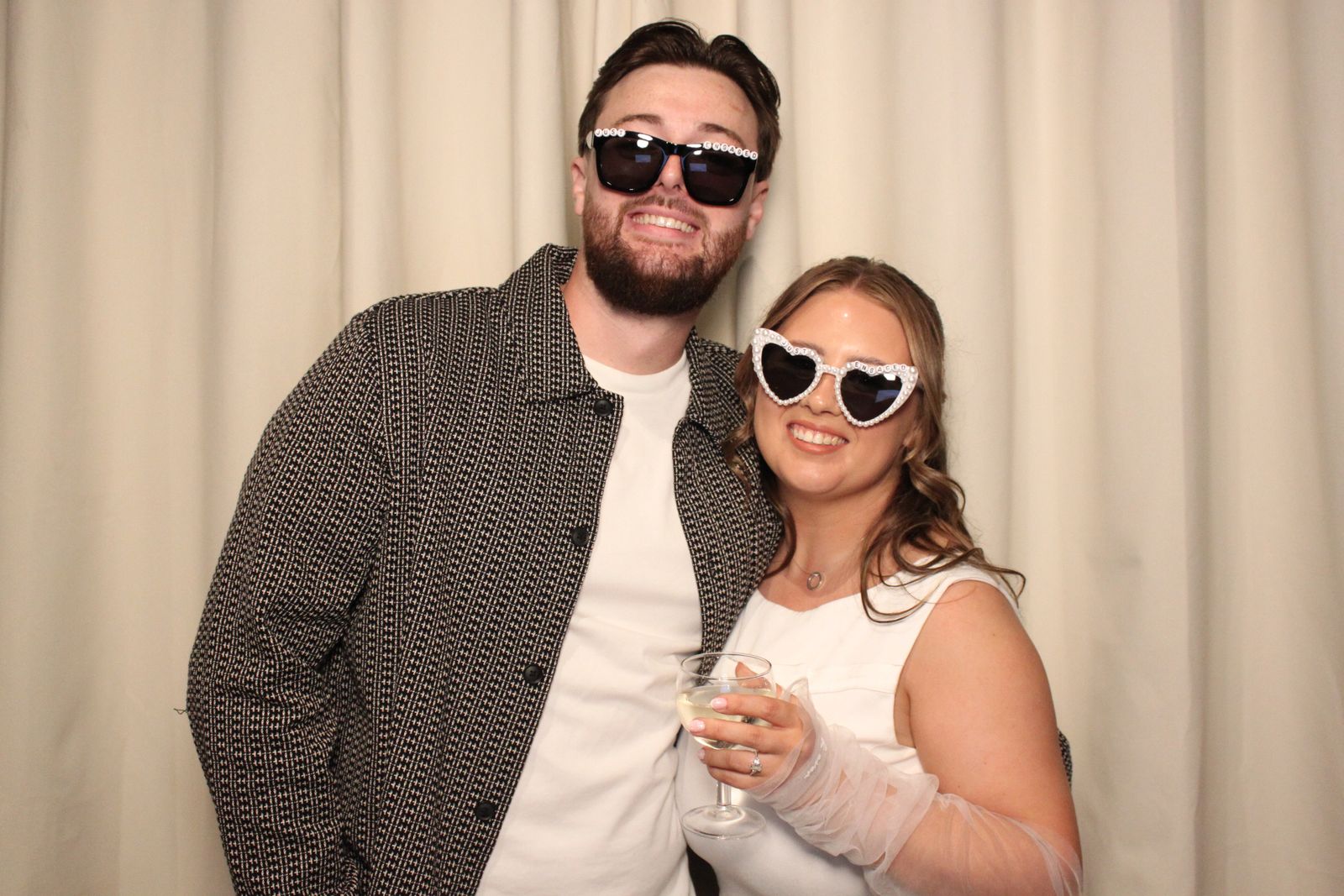 A smiling engaged couple in Just Engaged heart sunglasses with a glass of prosecco at the photo booth
