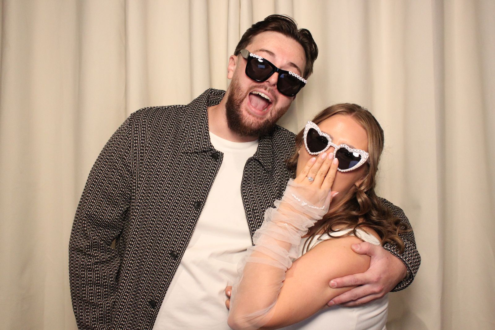 A surprised couple showing off an engagement ring with Just Engaged sunglasses at the photo booth