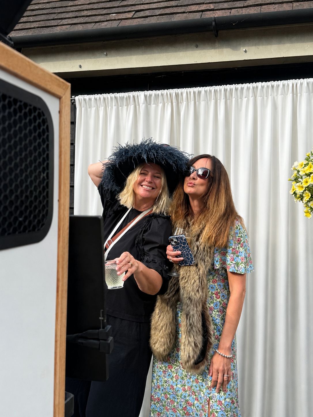 Two guests in feathered hats posing with drinks at the freestanding booth