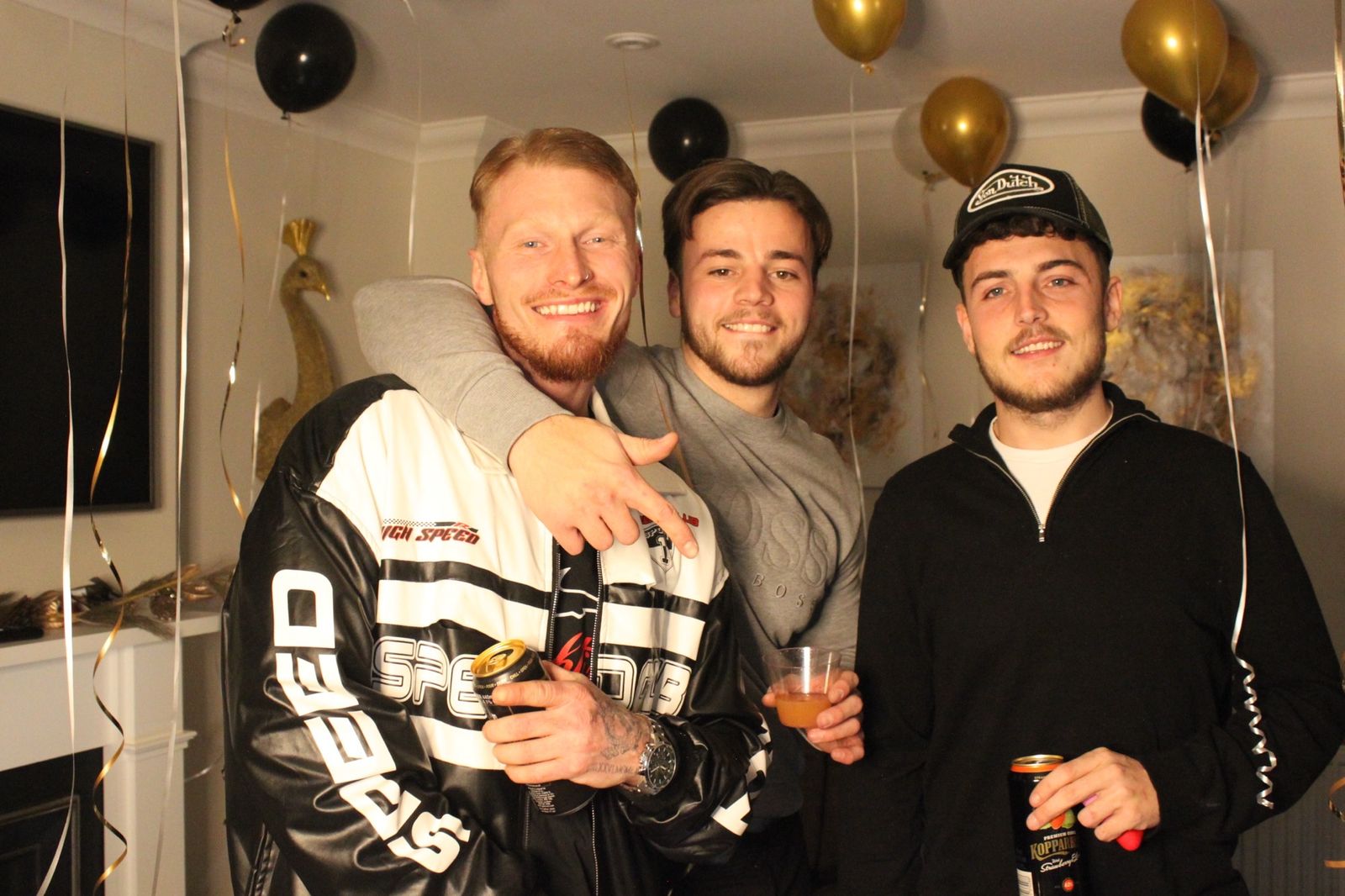 Three friends posing with drinks in front of black and gold NYE balloons