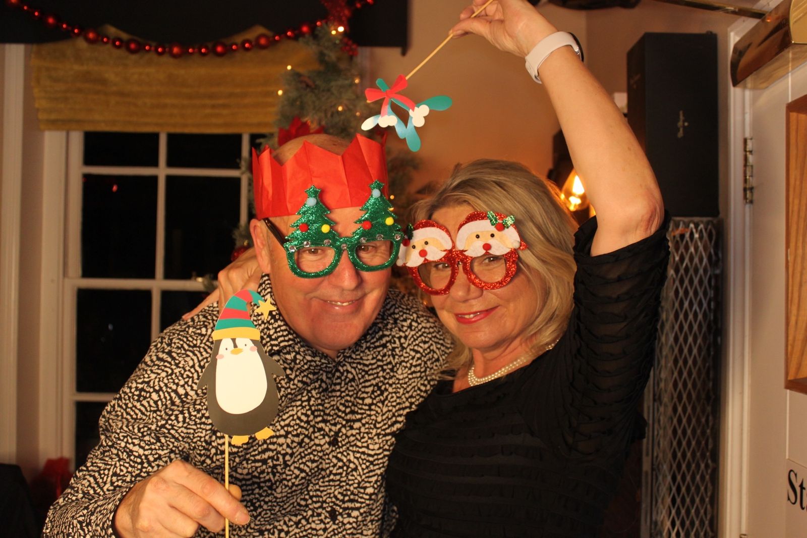 Couple in Christmas cracker crowns and festive glasses holding penguin and mistletoe props
